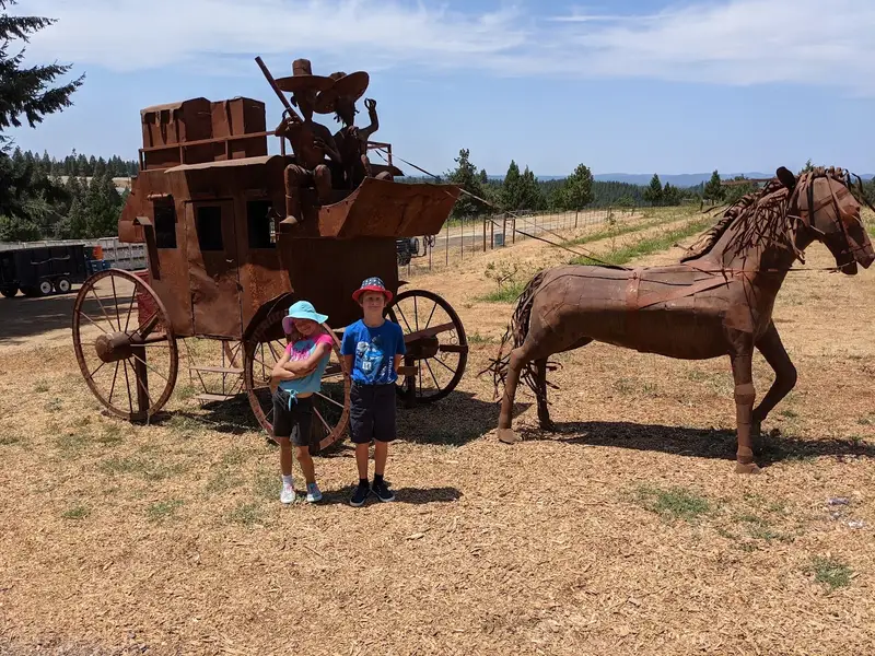 Two children on a family road trip posing beside a rustic stagecoach-and-horse sculpture in the sun — Suncoast Travel family and celebration travel for anniversaries, reunions, and multi-generational getaways